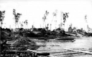 Black-and-white photograph of several log cabins on a small tree-lined hill. On the foreground there is a lake with a rudimentary dock as well as a few canoes.