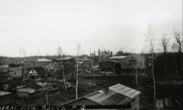 Black-and-white photograph of numerous log or board cabins and a barely cleared dirt road. There are trees standing in between the buildings.