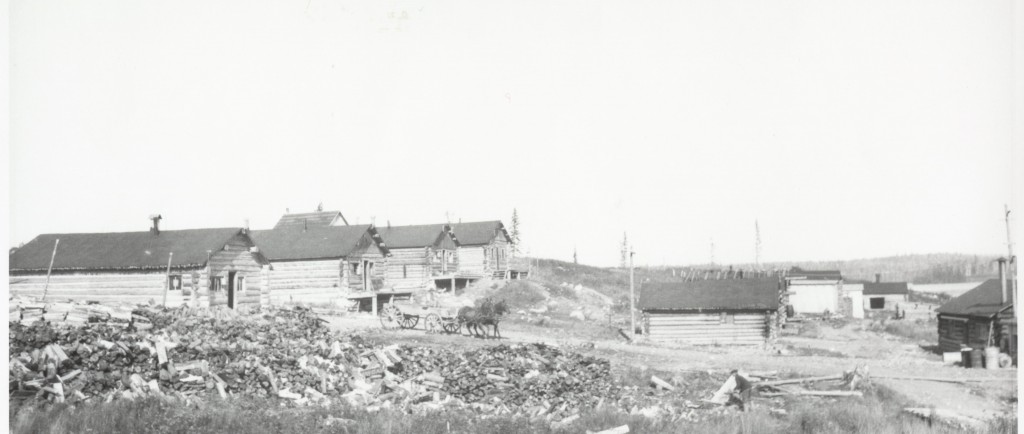 Photographie en noir et blanc de plusieurs cabanes en bois rond, d’une charrette tirée par deux chevaux et de nombreux amoncellements de bûches.