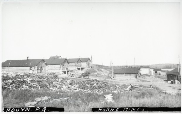 Black-and-white photograph of several log cabins, a horse-drawn wagon and many piles of logs.