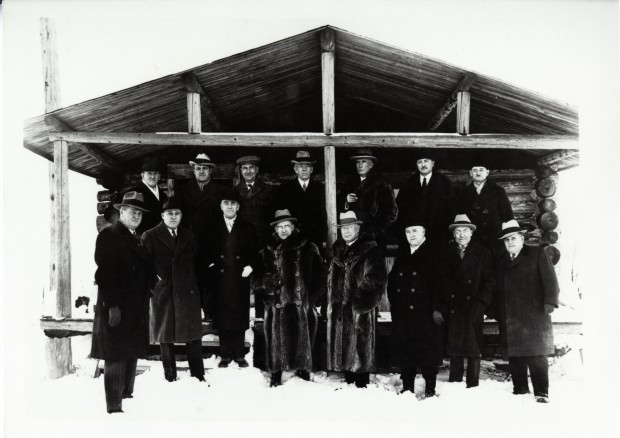 Black-and-white photograph of 15 men in fancy suits, three of whom are wearing fur coats, standing in front of a log cabin.