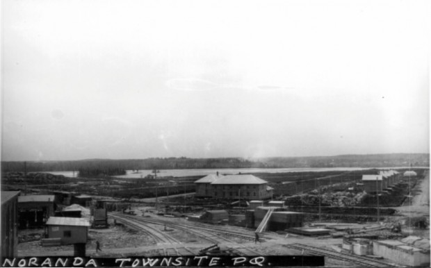 Black-and-white photograph of the construction site of the soon-to-be city of Noranda. You can see Osisko Lake in the background, the railroad in the foreground and the mine executives’ house on the right.