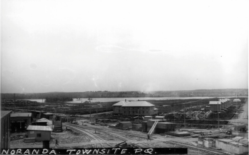 Black-and-white photograph of the construction site of the soon-to-be city of Noranda. You can see Osisko Lake in the background, the railroad in the foreground and the mine executives’ house on the right.