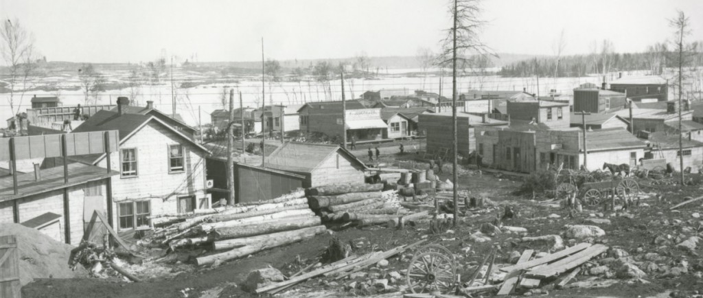 Photographie en noir et blanc de bâtiments rudimentaires construits au sud-ouest du lac Osisko. En avant-plan, des roches, des bûches, des planches et des débris.