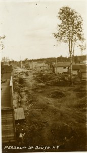 Photographie en noir et blanc de deux rangées de bâtiments rudimentaire, avec des trottoirs en bois et des arbres au centre.