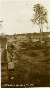 Black-and-white photograph of two rows of rudimentary buildings with boardwalks and trees in the middle.