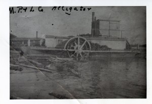 Photographie en noir et blanc, de mauvaise qualité, d’un bateau à vapeur qui est accosté. Sur la rive, un bâtiment et des billots flottent sur l’eau.