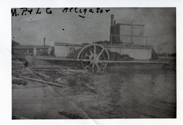Poor quality black-and-white photograph of a docked steamboat. On the shores, a building and logs are floating on the water.