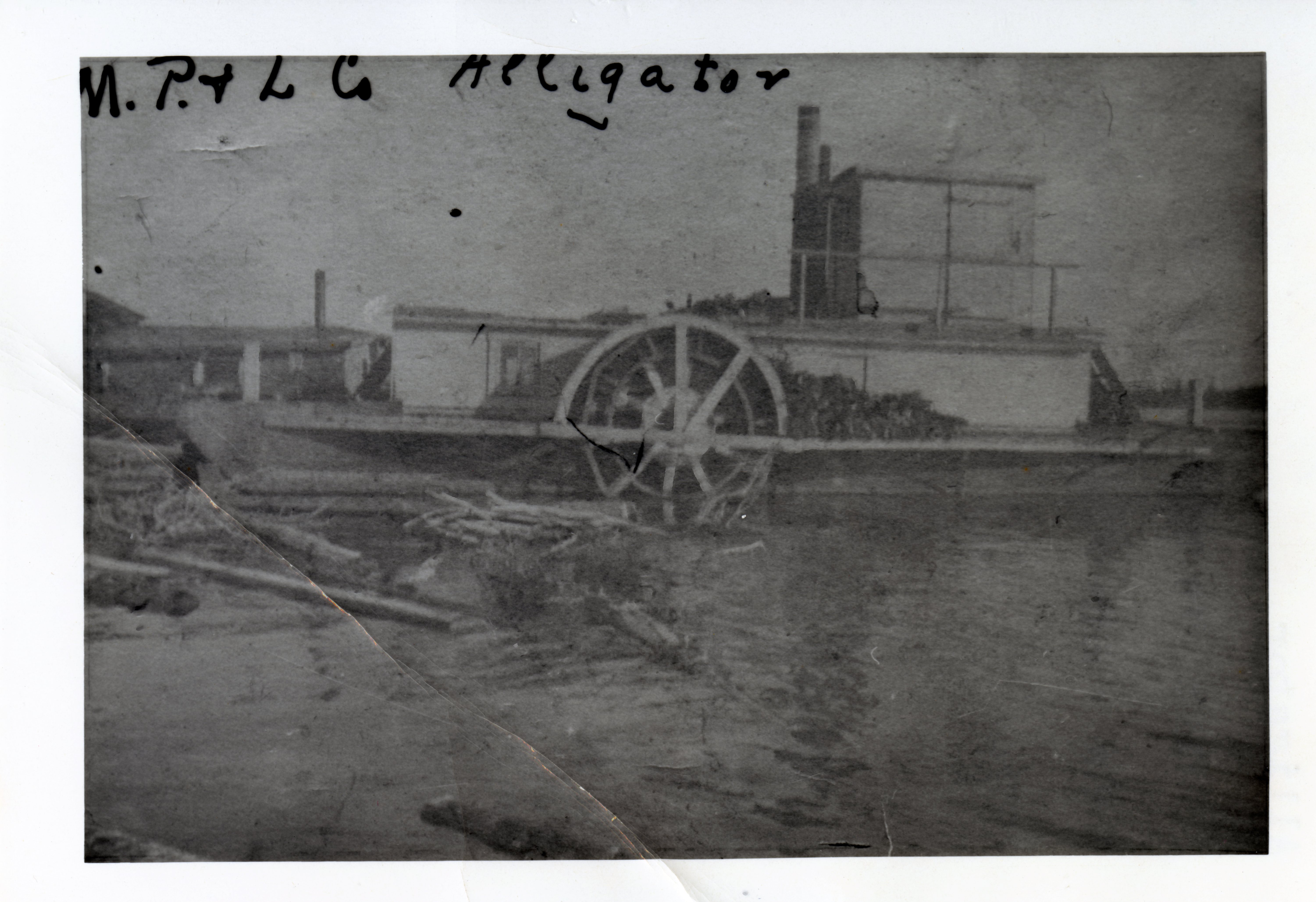 Poor quality black-and-white photograph of a docked steamboat. On the shores, a building and logs are floating on the water.