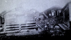 Poor quality black-and-white photograph of several rudimentary buildings. On the foreground you can see a log cabin.