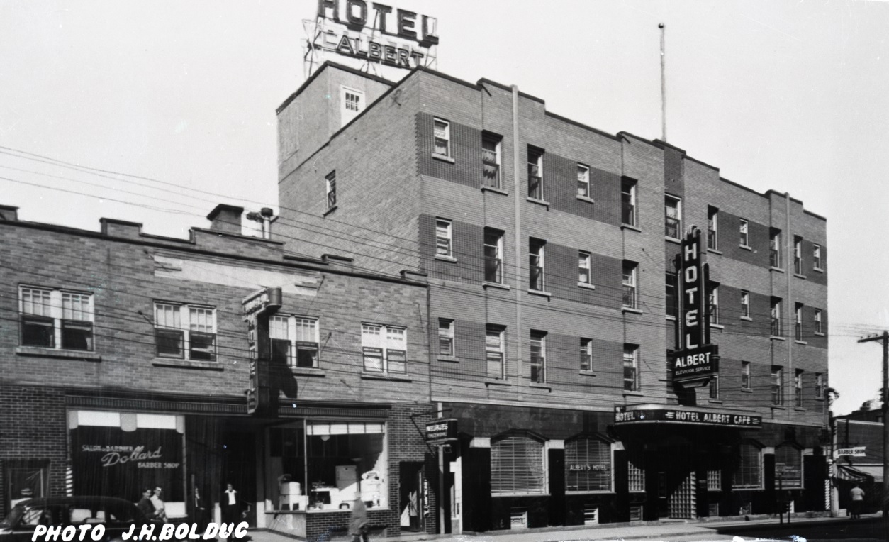 Black-and-white photograph of a four-story building with several signs, including one on the roof that reads, Hotel Albert. On the left, you can see a two-story building.