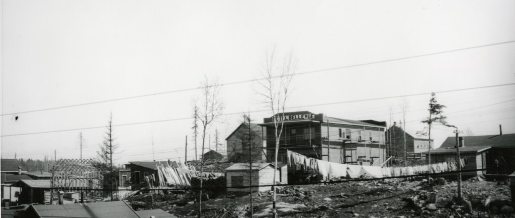 Photographie en noir et blanc de plusieurs bâtiments rudimentaires, en planche ou en bois rond, dont l’un avec l’inscription Hôtel Bellevue sur la façade de style Boomtowns.