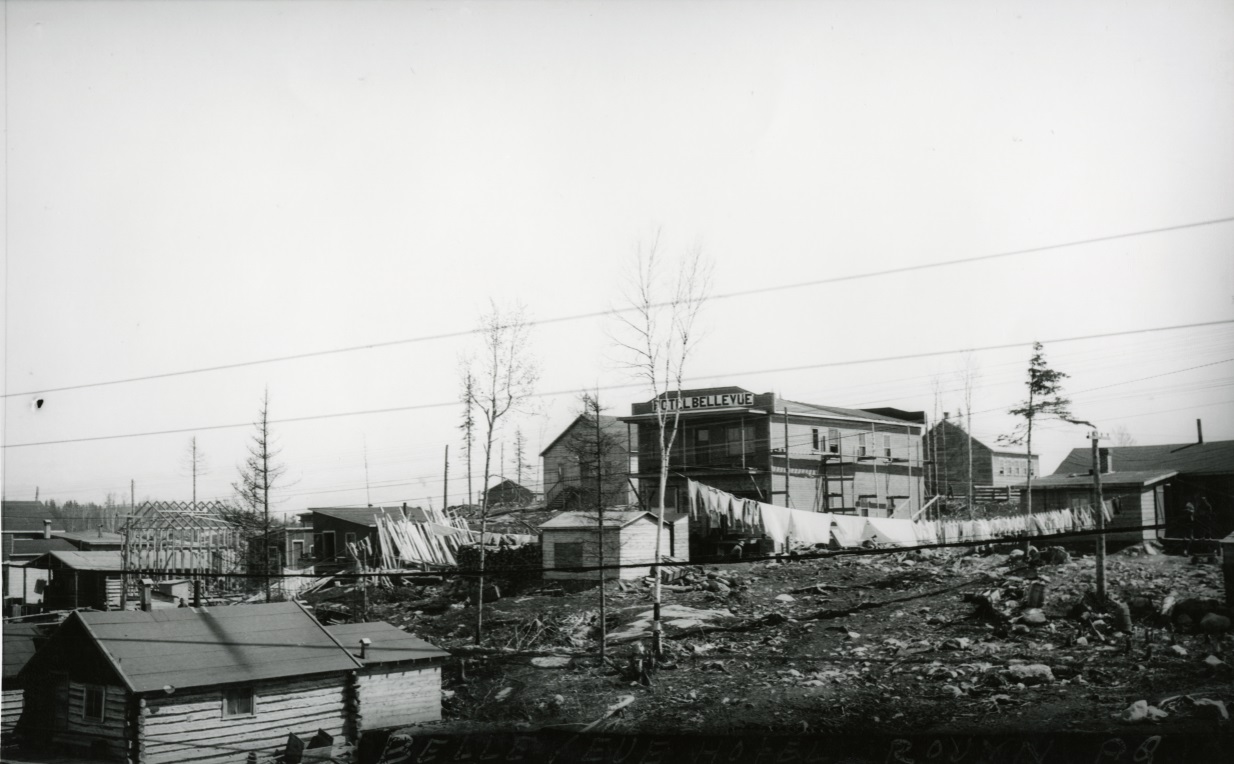 Black-and-white photograph of several rudimentary buildings made of planks or timbers. On one of the boomtown-styled fronts, you can read Hôtel Bellevue on a sign.