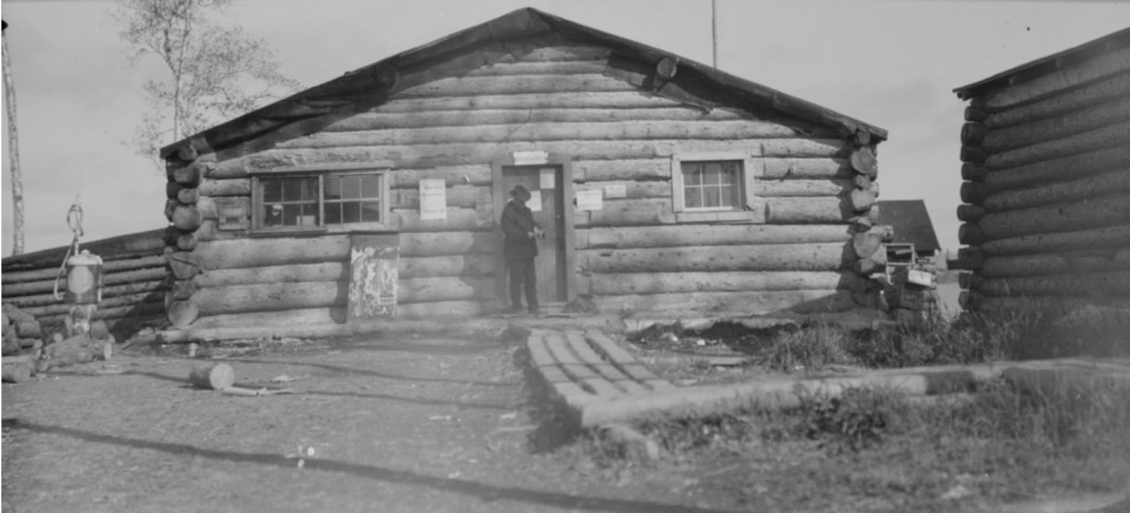 Photographie en noir et blanc d’un bâtiment en bois rond, avec un trottoir de bois menant à l’entrée. À l’avant, un homme en habit et une pompe à essence.