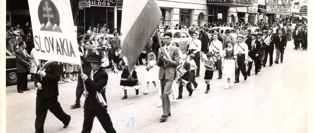 Black-and-white photograph of 25 people who parade before a crowd looking at them from the sidewalks. They’re marching in a parade with a sign that reads Slovakia decorated with the national coat of arms and a Slovakian flag.