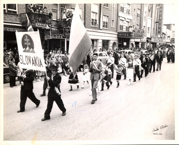 Black-and-white photograph of 25 people who parade before a crowd looking at them from the sidewalks. They’re marching in a parade with a sign that reads Slovakia decorated with the national coat of arms and a Slovakian flag.