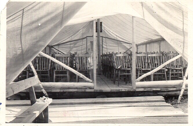 Photographie en noir et blanc de l’intérieur d’une tente  dans laquelle des chaises, disposées en rangées, sont installées sur un plancher de bois.