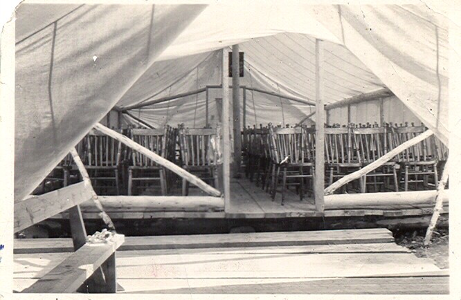 Black-and-white photograph of a tent interior showing rows of chairs and a hardwood floor.