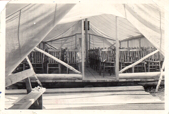 Black-and-white photograph of a tent interior showing rows of chairs and a hardwood floor.