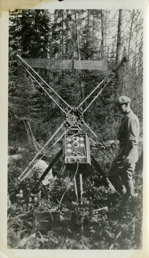 Photographie en noir et blanc d'un homme en habit de travail qui se tient debout à côté  du panneau de commande d'un appareil composé de quatre antennes disposé en croix. En arrière-plan, la forêt.