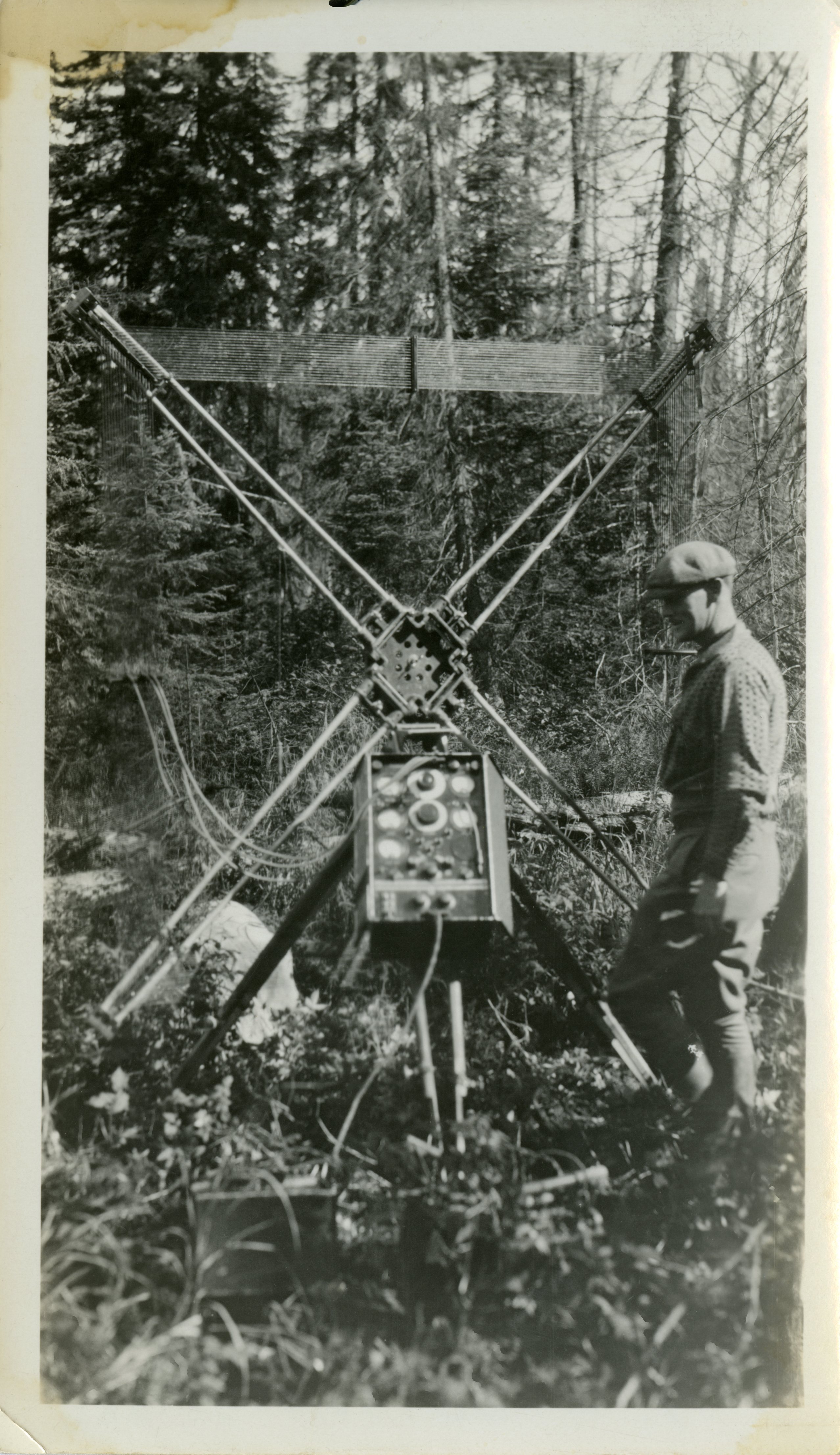 Black-and-white photograph of a man in work clothes standing beside the control panel of a device with four crossed antennas. A forest is visible in the background.