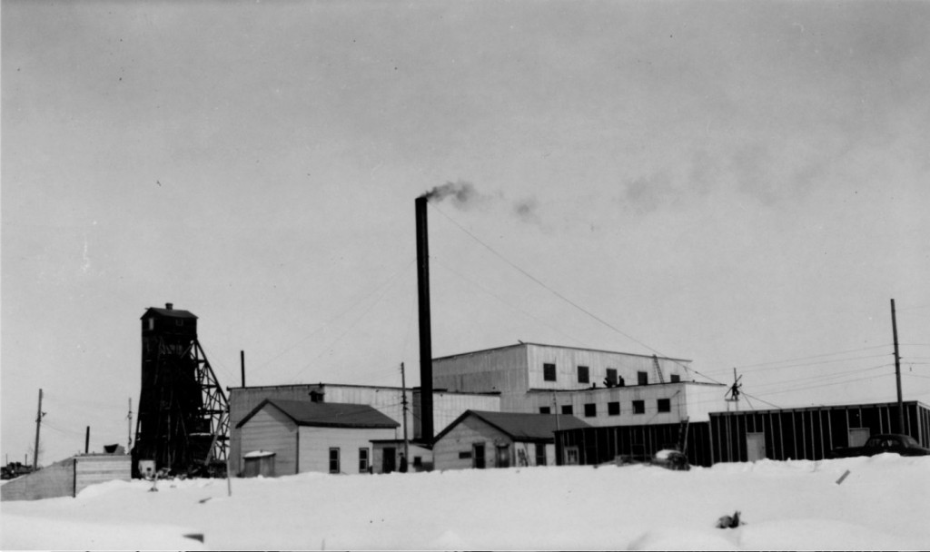 Black-and-white photograph of several mining buildings made of planks, including a headframe and a chimney and featuring a blanket of snow on the ground.