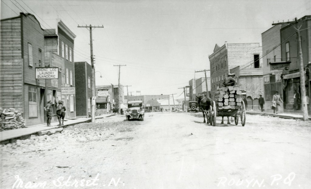 Black-and-white photograph of a gravel road where a car and a carriage are driving. People are walking on the boardwalks. The buildings have boomtown-styled fronts. A sign reads, Chong Lee Laundry Buanderie.