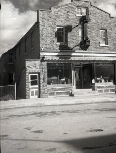 Photographie en noir et blanc d'un édifice de trois étages en brique. En plein milieu de l'édifice, une affiche annonce le Princess Hotel et à gauche, une porte semble menée au sous-sol.