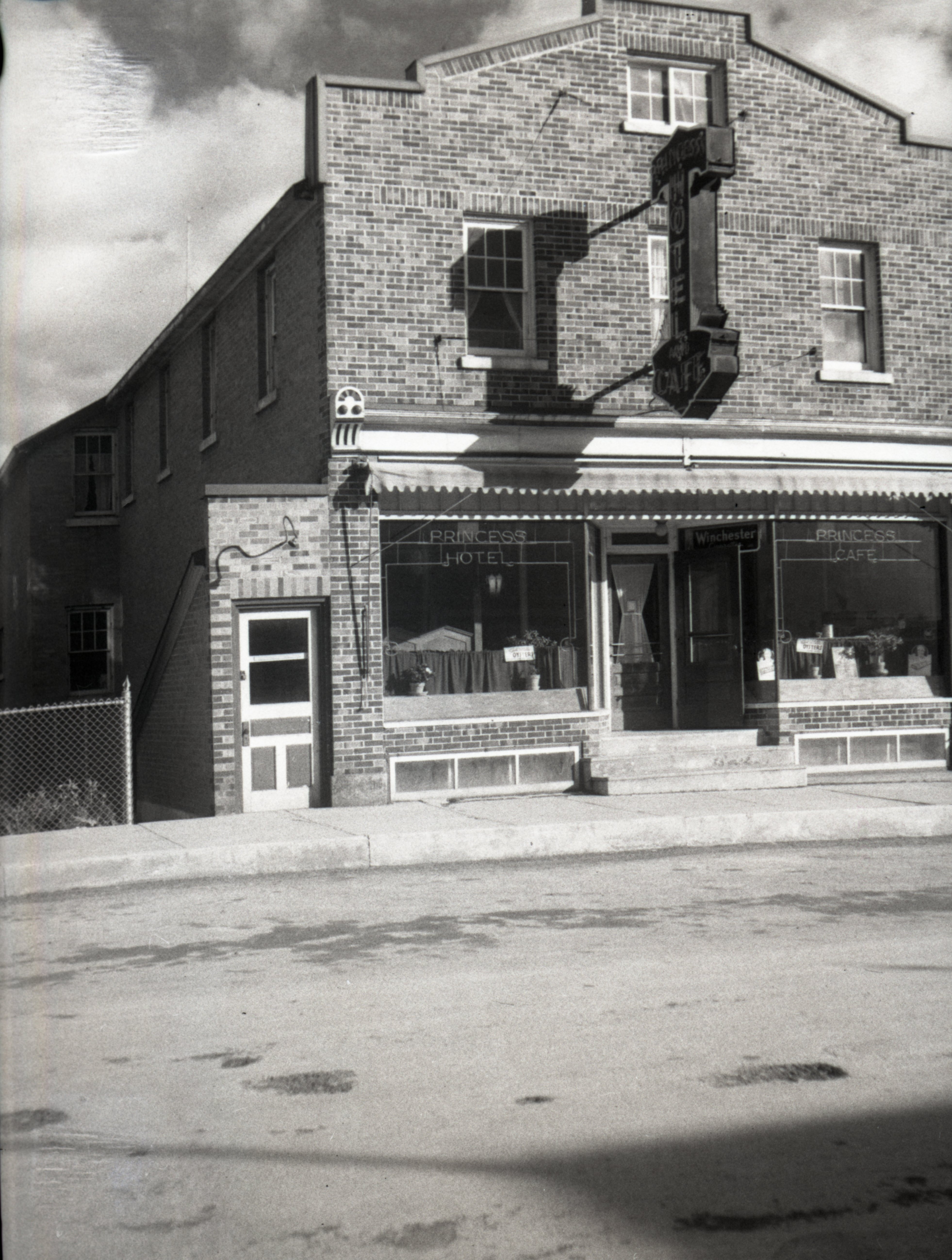Black-and-white photograph of a three-story brick building. On the middle of its front, you can read Princess Hotel and on the left, you can see a door apparently leading to a basement.