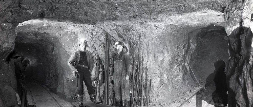 Photographie en noir et blanc de l’intersection entre trois galeries à l’intérieur d’une mine. Il y a deux rails de chemin de fer par terre, ainsi que deux hommes qui posent devant une foreuse et des aciers.