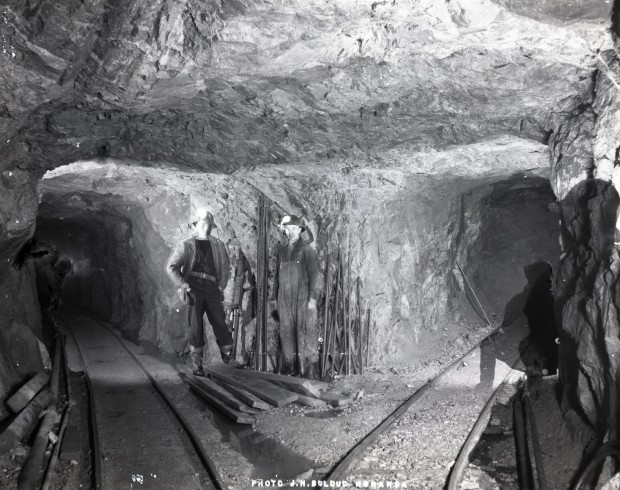 Black-and-white photograph of the intersection of three roads inside a mine. There are two railroad tracks on the ground and two men pose in front a drill machine and a steel structure.