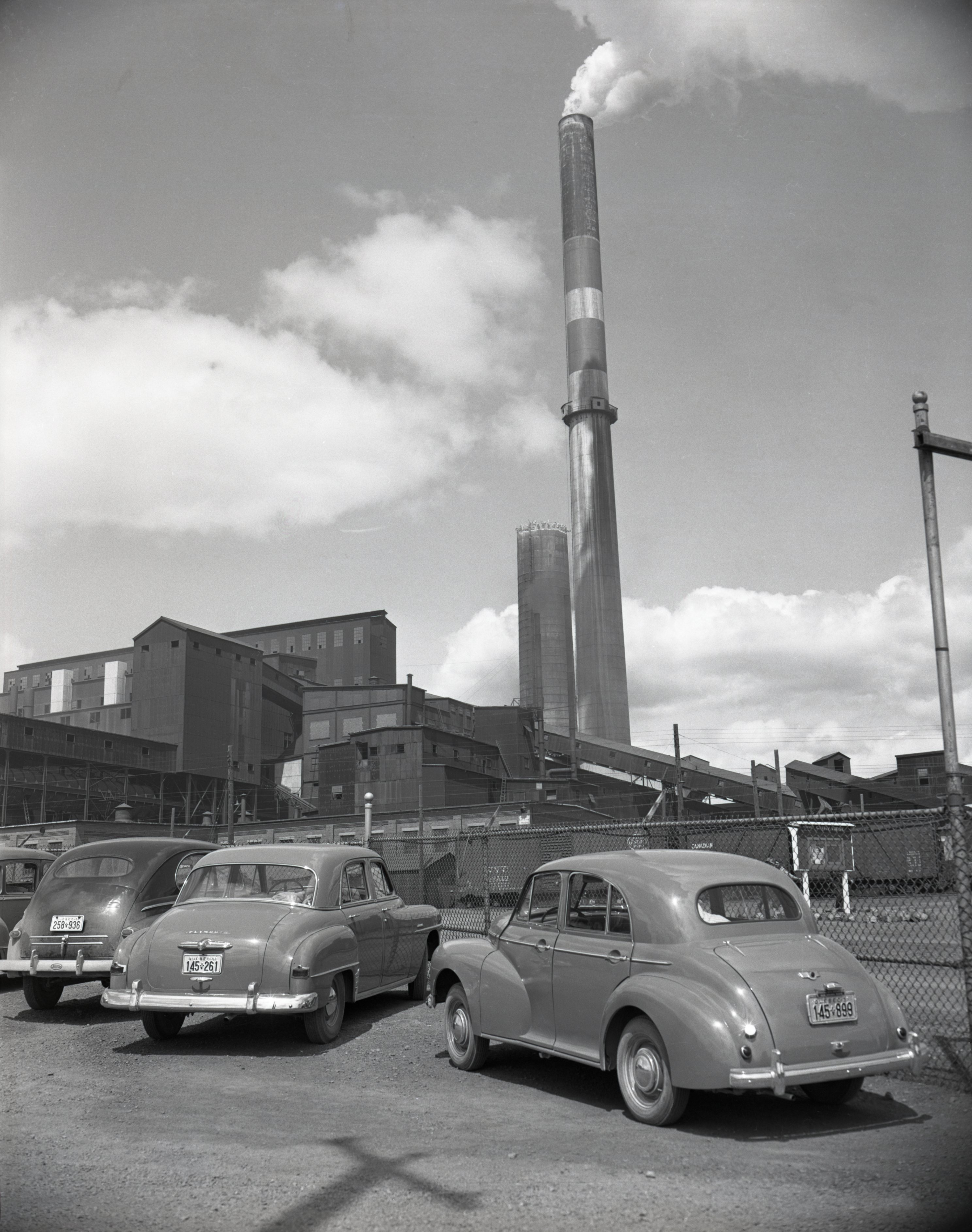 Black-and-white photograph of three old cars parked in front of the Horne mine’s fence. You can see the smelter with smoke rising from a chimney in the background.