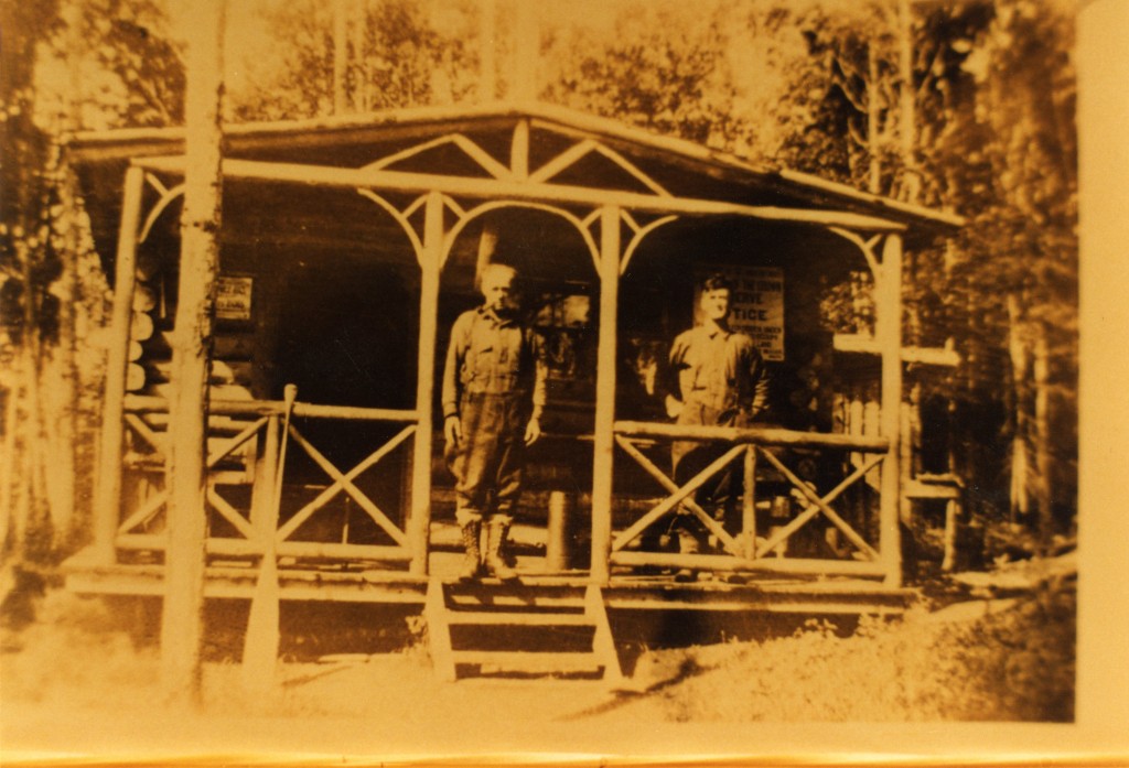 A sepia-toned photograph of two men standing on the porch of a log cabin with a forest in the background.