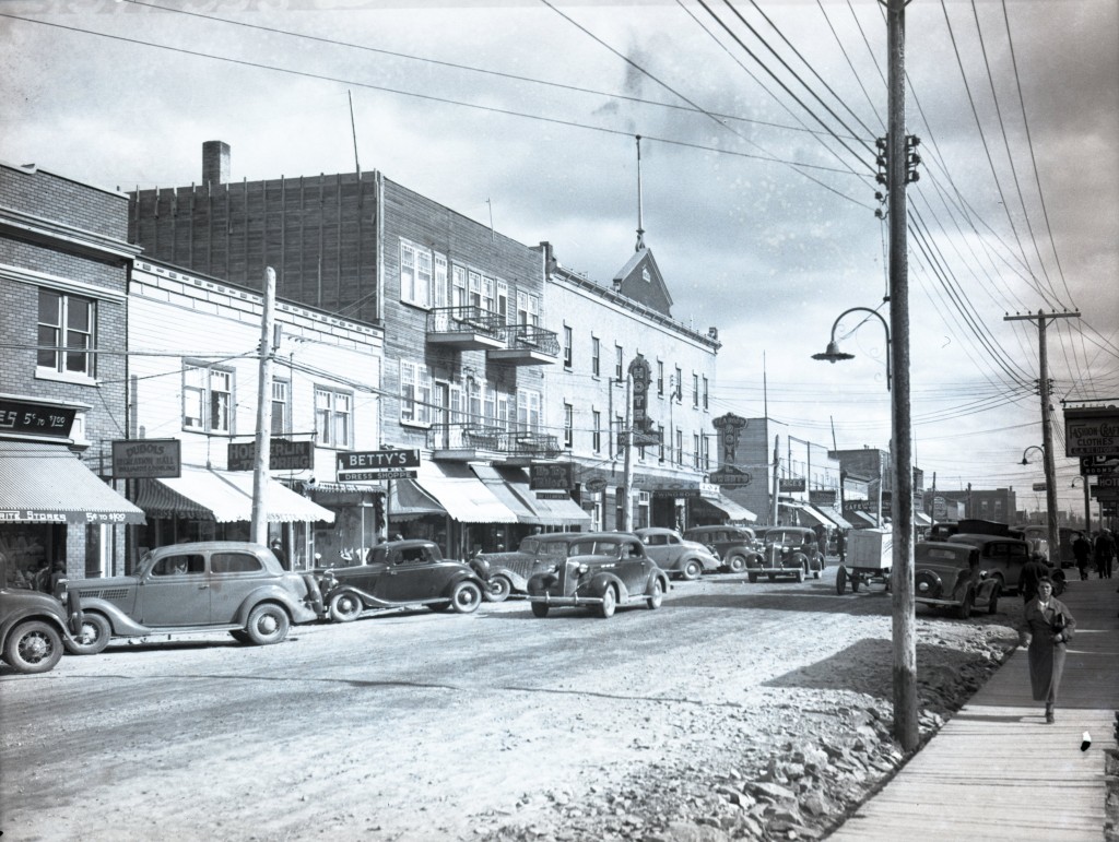 Black-and-white photograph of a street lined with two or three-story buildings on which there are signs for some of the stores’ name. You can see several people walking on the boardwalks as well as cars that are either parked or rolling.