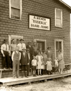 Photographie noir et blanc de seize personnes, dont plusieurs femmes et enfants, devant un bâtiment dont une enseigne indique  A. Niemen Ruokala Board & Rooms.