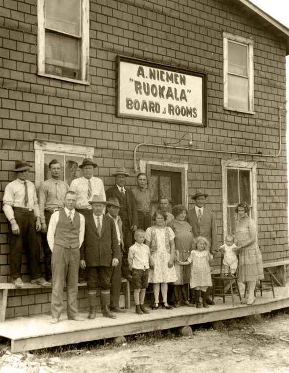 Black-and-white photograph of several women and children before a building whose sign reads, A. Niemen Ruckala Board & Rooms.