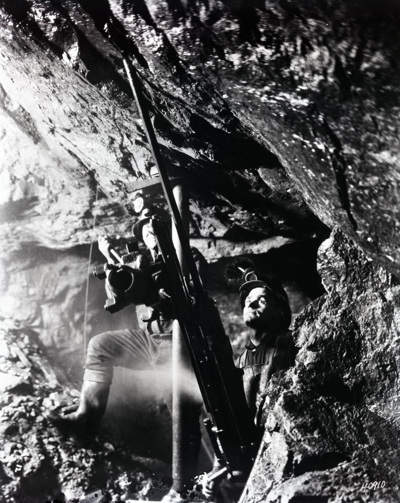 Photographie noir et blanc de deux mineurs qui opèrent une foreuses fixée au sol et au plafond avec des tiges de métal. Ils ont des bottes de caoutchous et un casque avec lumière.