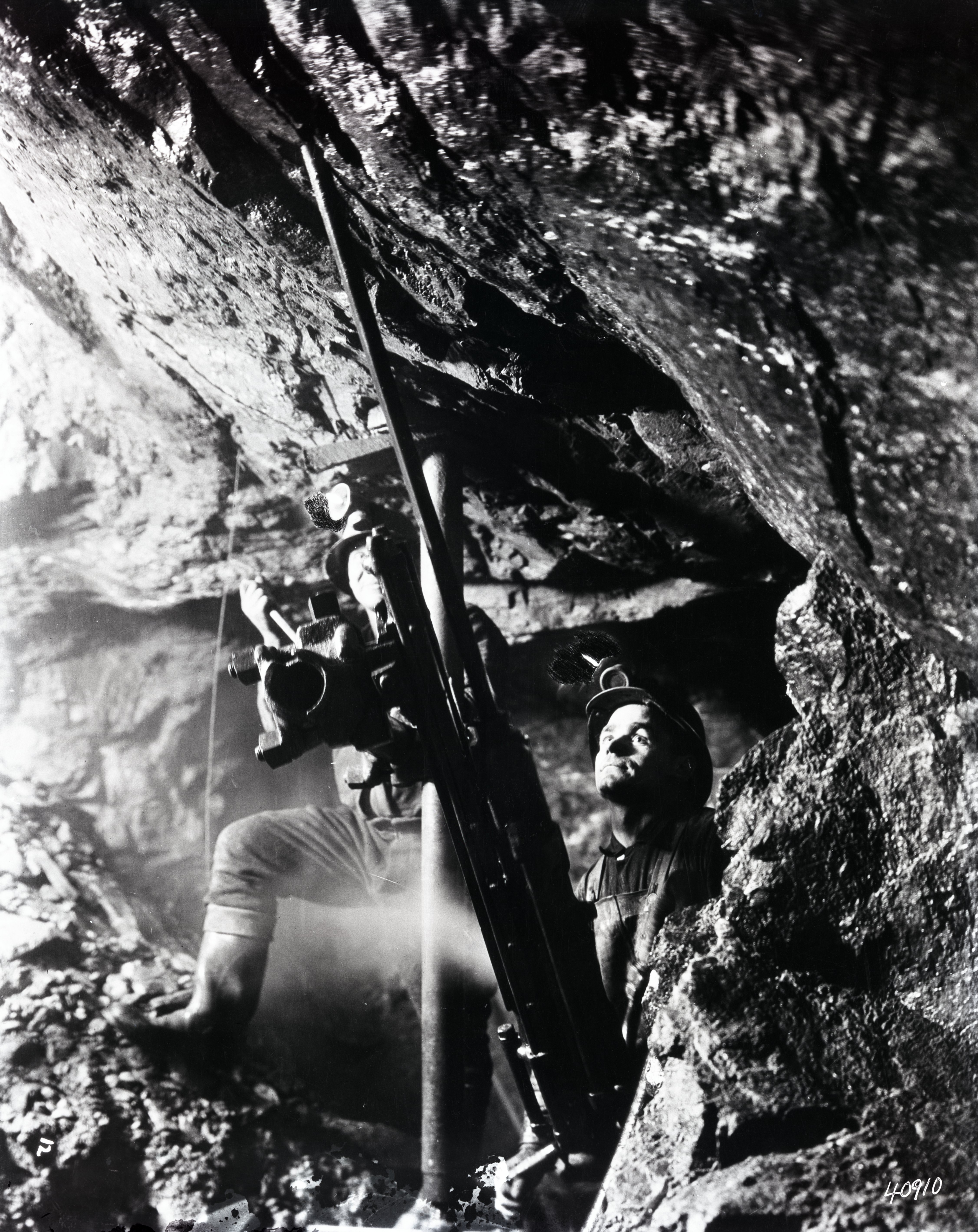 Black-and-white photograph of two miners operating a drill mounted to the ground and roof with metal rods. The men wear rubber boots and a helmet with a light.