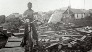 Black-and-white photograph of a little boy on a bicycle, posing in front of a pile of planks and logs. In the background, you can see houses and a church.