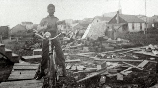 Black-and-white photograph of a little boy on a bicycle, posing in front of a pile of planks and logs. In the background, you can see houses and a church.