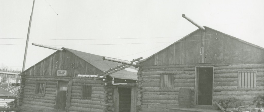 Black-and-white photograph of two log cabins with bars on the windows. A sign is put up on the door of the left building.