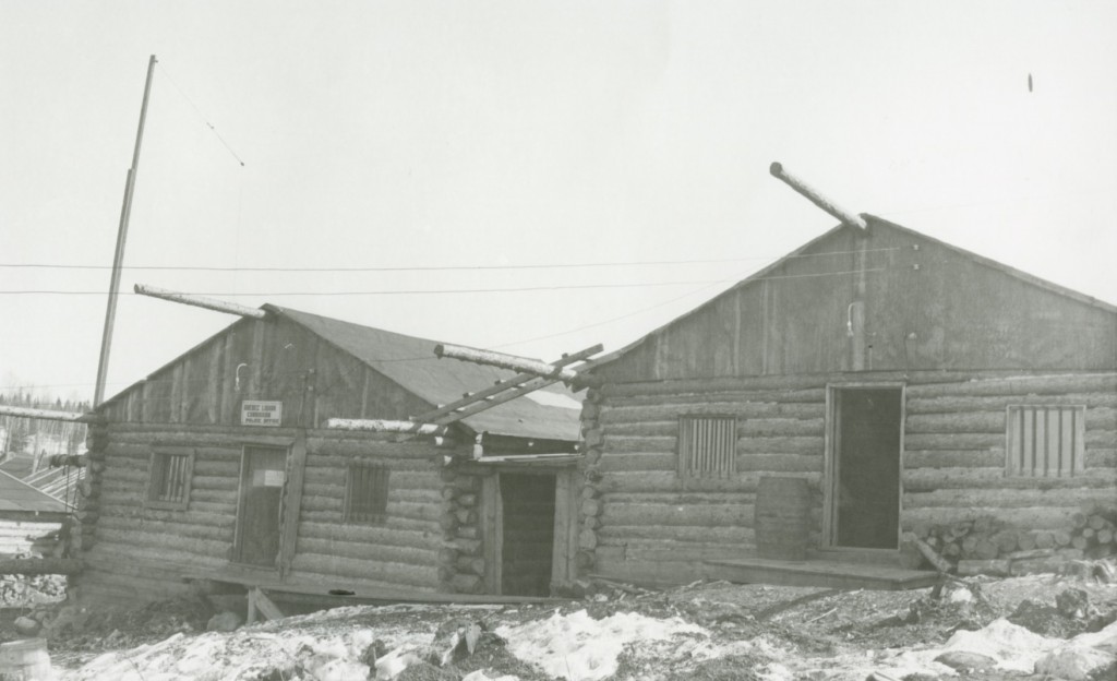 Black-and-white photograph of two log cabins with bars on the windows. A sign is put up on the door of the left building.
