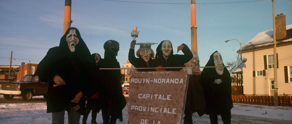 Photo couleur d'un groupe de sept personnes, devant la fonderie Horne, qui sont déguisé et qui transporte une affiche qui porte l'inscription : Rouyn-Noranda capitale provinciale de la pollution.