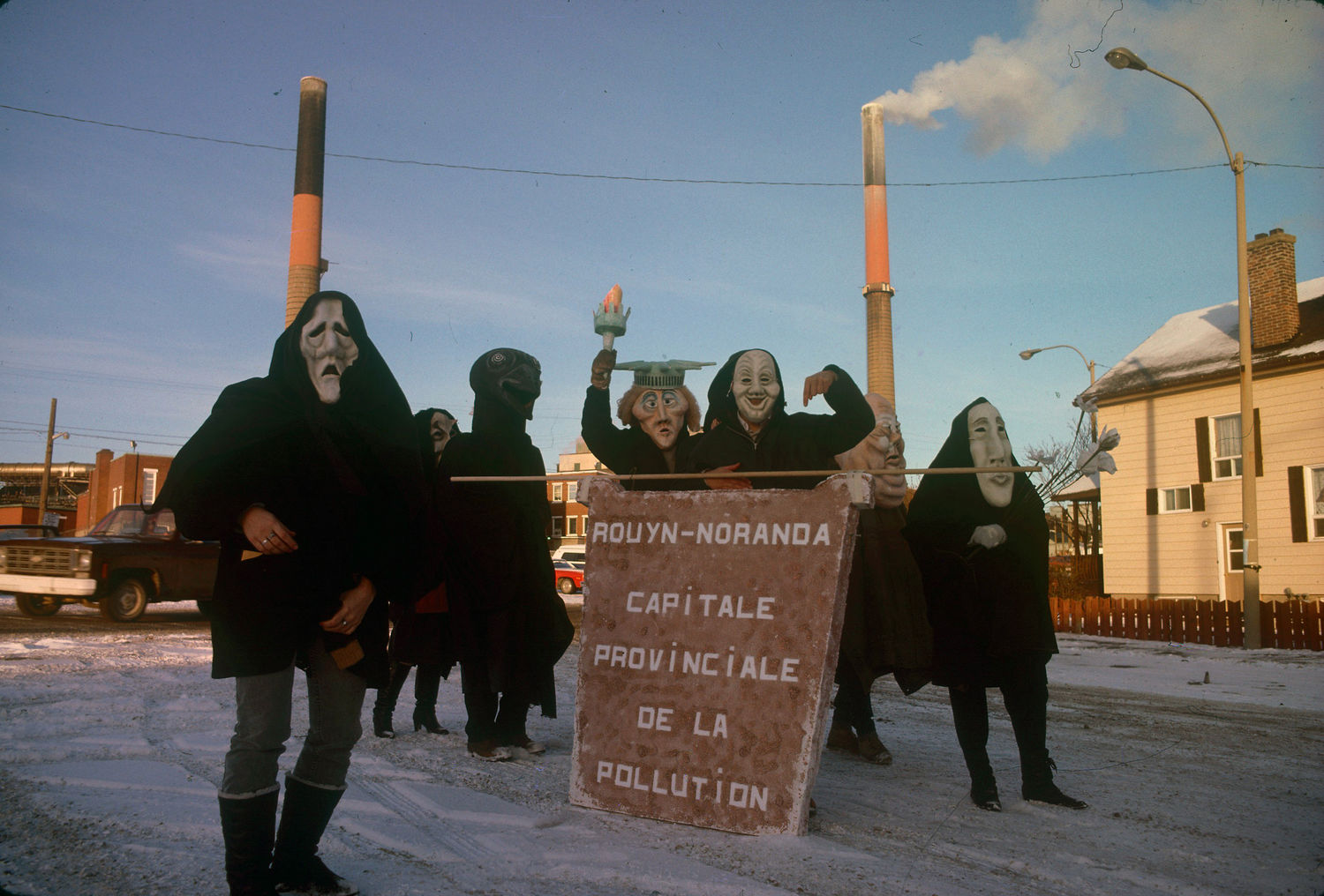 Colour photograph of seven people in front of the Horne smelter. They are costumed and carry a poster that reads in French Rouyn-Noranda provincial capital of pollution.