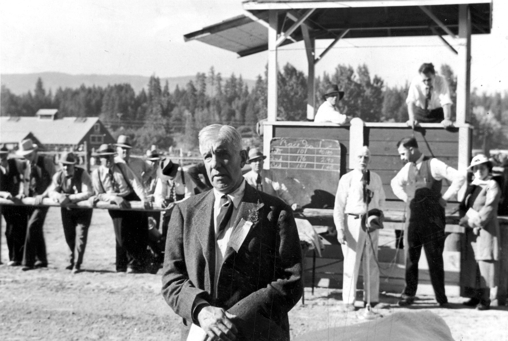 Black and white photo of an older man wearing a suit, other men standing and an announcement stand are in the rear.