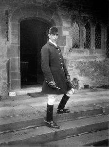 Black and white photo of a young man wearing riding boots and jacket, standing on the steps of a stone building.