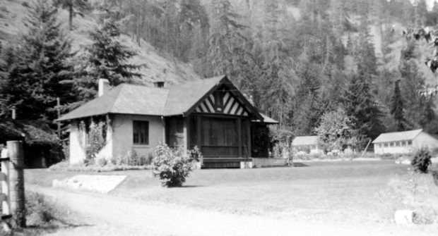 Black and white photo of a gabled house, two smaller buildings to the right, hill in background.