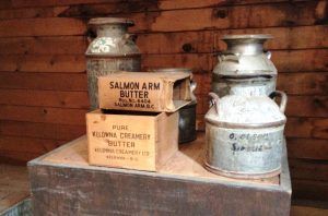 Colour photo of four milk cans, two butter boxes on a wooden stand.