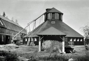Black and white photo of a round wooden barn with machinery attached to the side, waste fodder in front.