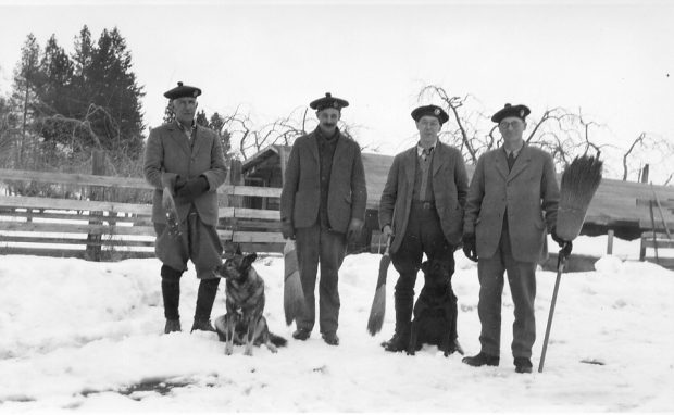 Black and white photo of four men standing in the snow holding brooms. The men are all wearing tams and suit jackets. A dog is seated with them.